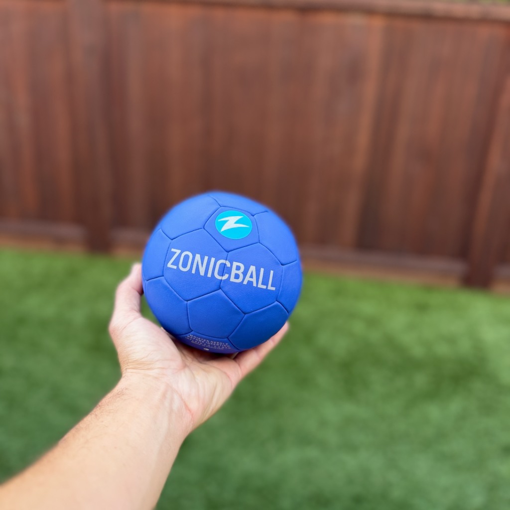 A hand holding a Zonicball mini soccer ball in an outdoor backyard setting with a wooden fence and green grass in the background.