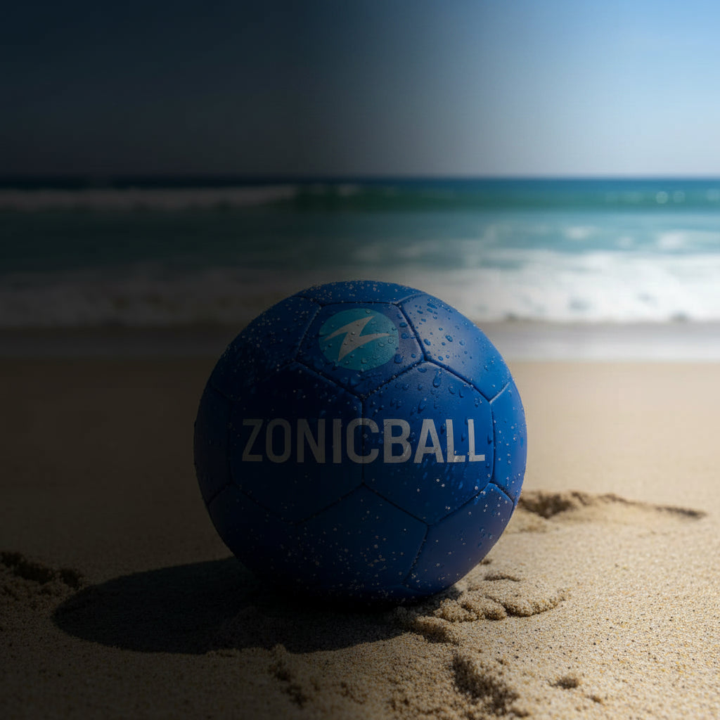 A blue Zonicball soccer ball with white lettering sits on a sandy beach with the ocean and blue sky blurred in the background.